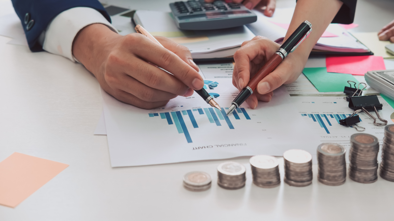 Una mano de una mujer y un hombre sostienen sendas plumas que señalan un gráfico y tienen delante monedasa